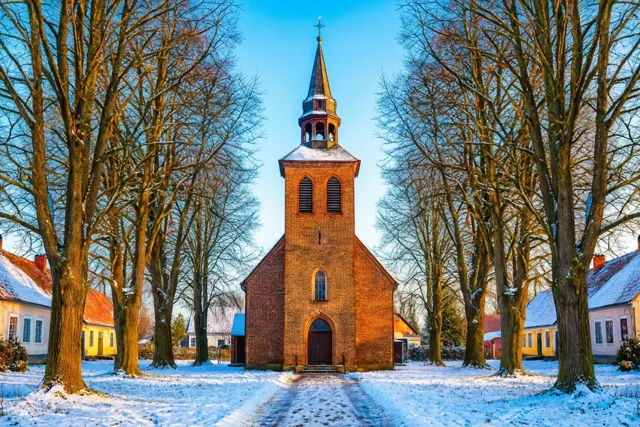 Een kleine bakstenen kerk met een spitse toren staat op een besneeuwd kerkhof tussen kale bomen in goudkleurig licht onder een blauwe hemel.