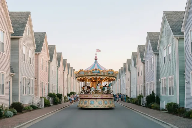 Een kleurrijke draaimolenwagen rijdt door een straat met pastelkleurige trapgevelhuizen in zacht, gelijkmatig licht.