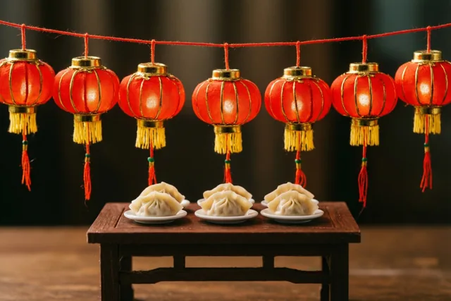 Een rij miniatuurlantaarns in het rood hangt boven een kleine houten tafel met drie bordjes kleine dumplings, in warm licht met rode en gouden tinten.