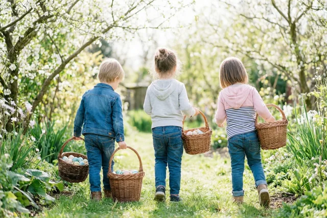 Drie kinderen lopen van achteren gezien met rieten mandjes gekleurde eieren door een zonnige tuin onder witbloeiende bomen.