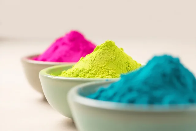 Drie kleine kommen met roze, geelgroen en blauw poeder staan schuin achter elkaar op een effen lichte ondergrond.