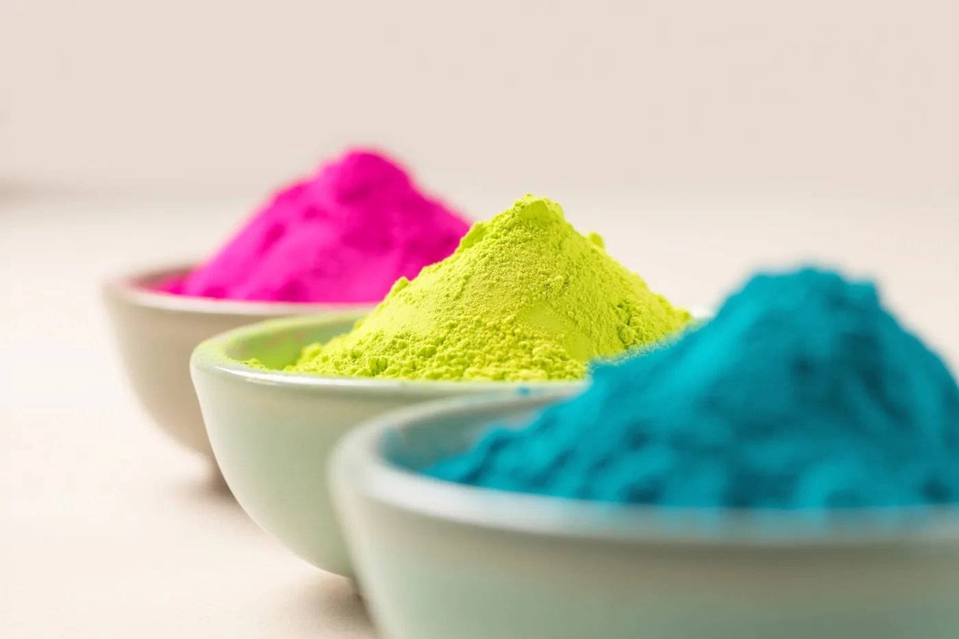 Drie kleine kommen met roze, geelgroen en blauw poeder staan schuin achter elkaar op een effen lichte ondergrond.