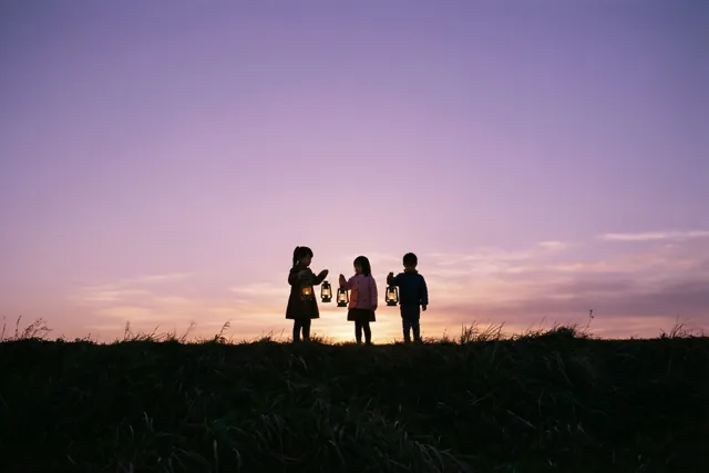 Drie kleine kinderen staan als silhouetten op een met gras begroeide dijk, elk met een brandende lantaarn tegen een roze en paarse schemerhemel.