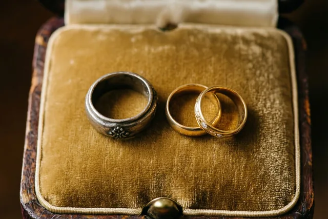 Een zilveren en twee gouden ringen liggen op een versleten goudbruin fluwelen kussen in een antiek ringendoosje.
