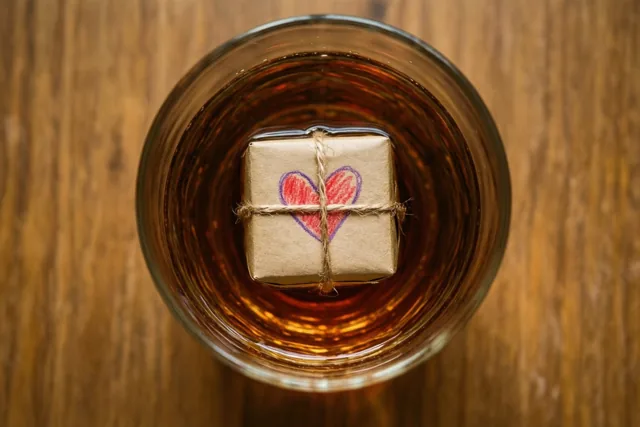 Een klein cadeau in bruin papier met touw en een door een kind met waskrijt getekend hart drijft in een glas amberkleurige cognac op een warme houten ondergrond.