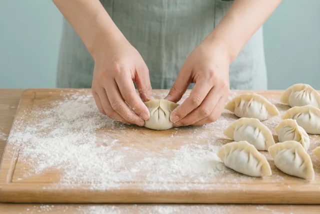 Handen vouwen dumplings op een met bloem bestoven houten plank, met een groene schort en een lichtblauwe achtergrond in beeld.