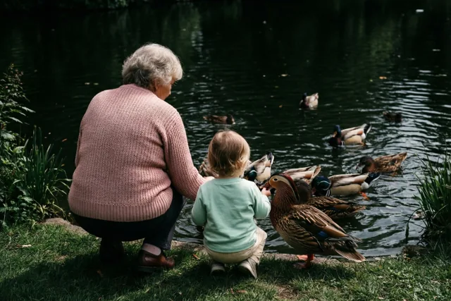 Een oudere vrouw en een klein kind hurken bij een vijver om eenden te voeren, met groen gras langs het donkere water.