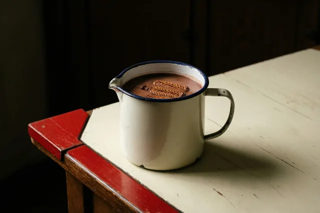 Een witte emaille kan chocolademelk met twee speculaasjes staat op een verweerde witte houten tafel met een rode mijter, tegen een donkere houten achtergrond.