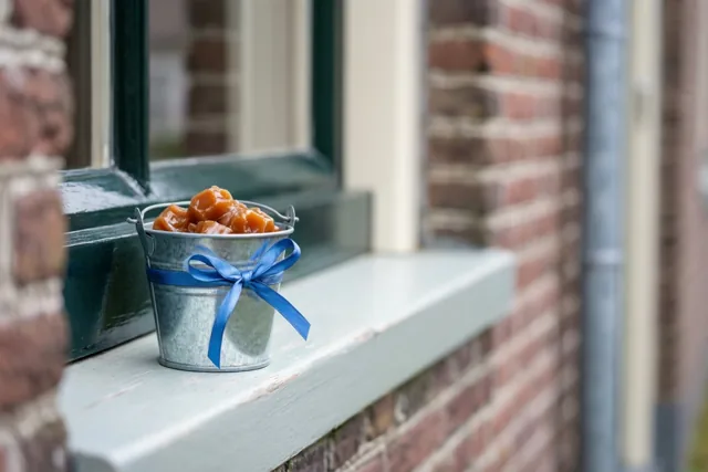Een verzinkt metalen emmertje met karamelsnoepjes en een blauw lint staat op een lichtgroene vensterbank tegen een rode bakstenen muur met een donkergroen raamkozijn.