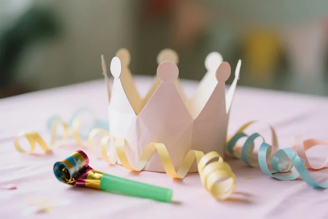Een papieren feestkroon ligt met kleurrijke krulslingers en een roltong op een pastelroze tafelkleed in zacht licht.