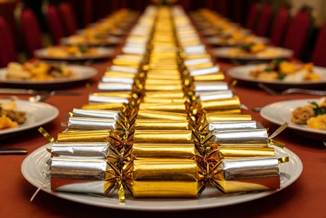 Een lange tafel met een rood tafelkleed en rode stoelen is gedekt met witte borden waarop gouden en zilveren feestcrackers liggen.