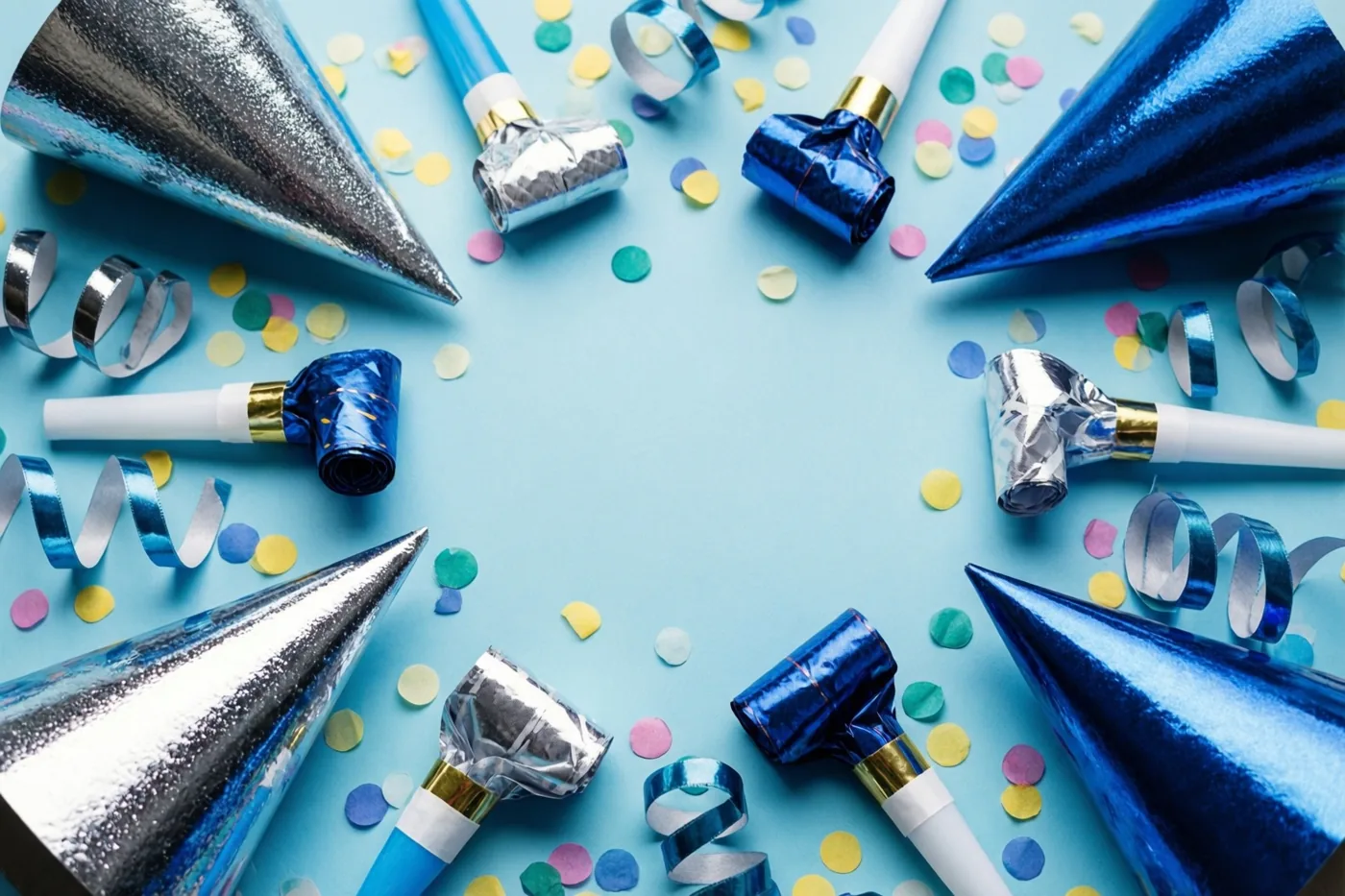 Feesthoedjes, roltongen, confetti en gekrulde linten liggen van bovenaf gezien in een cirkel op een pastelblauwe ondergrond, in blauwe, zilveren en pasteltinten.