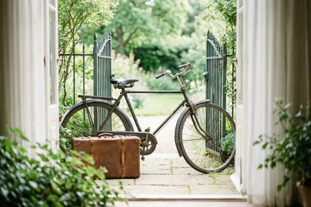 Door een open deuropening is een oude fiets met een bruine leren koffer te zien tegen een groen metalen tuinhek in een weelderige tuin bij zacht natuurlijk licht.