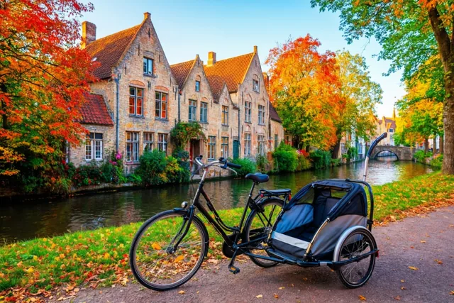 Een zwarte fiets met een kinderkar staat langs een kanaal bij oude gevelhuizen, omringd door rood en oranje herfstblad onder een helderblauwe lucht.