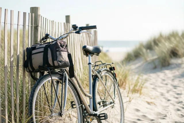 Een fiets met een strandtas op de bagagedrager leunt tegen een houten duinhek op een zandstrand, in helder daglicht met een zacht nevelige achtergrond.