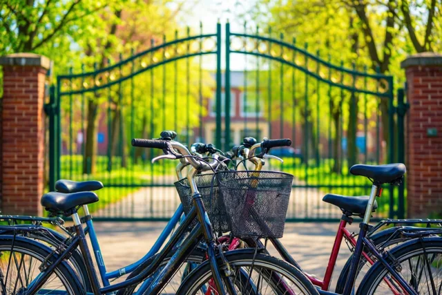 Fietsen met manden staan voor een groene metalen boogpoort tussen rode bakstenen pijlers en groen blad op een zonnige lentemorgen.