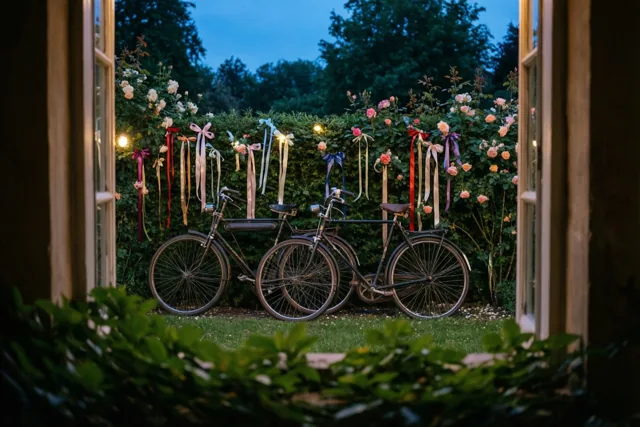 Door een open raam zijn twee oude fietsen te zien die tegen elkaar leunen bij een haag met rozen en kleurrijke linten, in de schemering verlicht door warme lichtslingers.