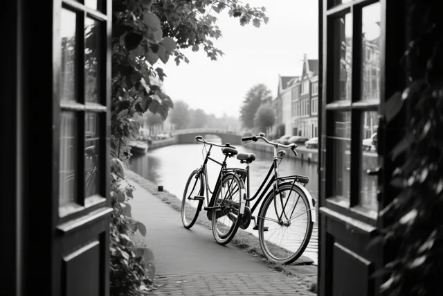 Door een open deuropening zijn twee fietsen te zien die tegen elkaar leunen op een verhard pad langs een kanaal, in zwart-wit.