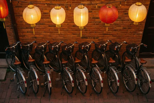 Een rij fietsen met manden en bagagedragers staat tegen een rode bakstenen muur, met warm geel en rood gloeiende papieren lampionnen erboven.