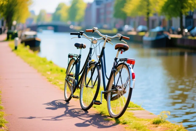 Twee fietsen leunen tegen elkaar op een rood bakstenen pad langs een kanaal, met groen gras en bomen in warm zonlicht.