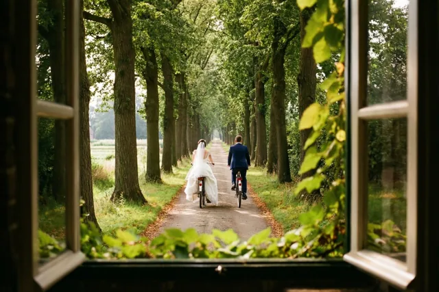 Door een open raam met groene bladeren op de voorgrond is een bruid in een witte jurk te zien die naast een bruidegom in een blauw pak door een bomenlaan fietst.