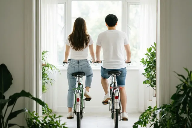 Twee mensen fietsen binnenshuis naar een groot raam, van achteren gezien in een lichte ruimte met witte muren en groene planten.