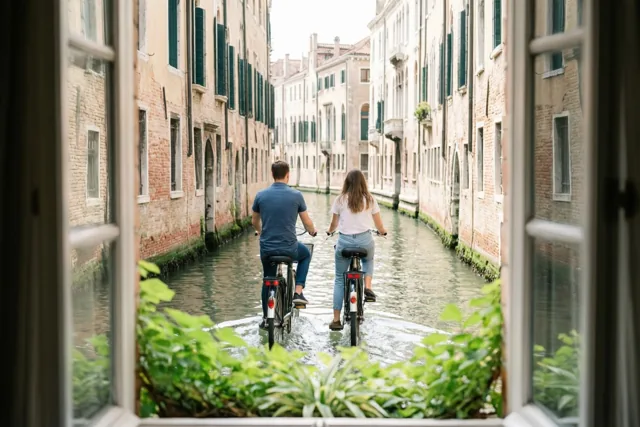 Door een raamkozijn met groen op de voorgrond zijn twee vrienden van achteren te zien die langs een smal kanaal tussen oude verweerde gebouwen fietsen.