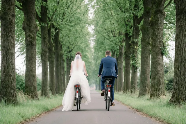 Een bruid in een witte trouwjurk en een bruidegom in een blauw pak fietsen naast elkaar over een verharde laan met hoge bomen op regelmatige afstand, gezien van achteren.