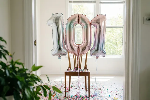 Drie folieballonnen vormen 101 en zijn aan een houten stoel vastgemaakt in een lichte kamer met kleurrijke confetti op de vloer en een raam achteraan.