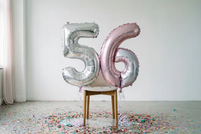 Twee grote folieballonnen in de vorm van 5 en 6 hangen aan een houten stoel in een lichte kamer met kleurrijke confetti en zilveren, roze en witte tinten.