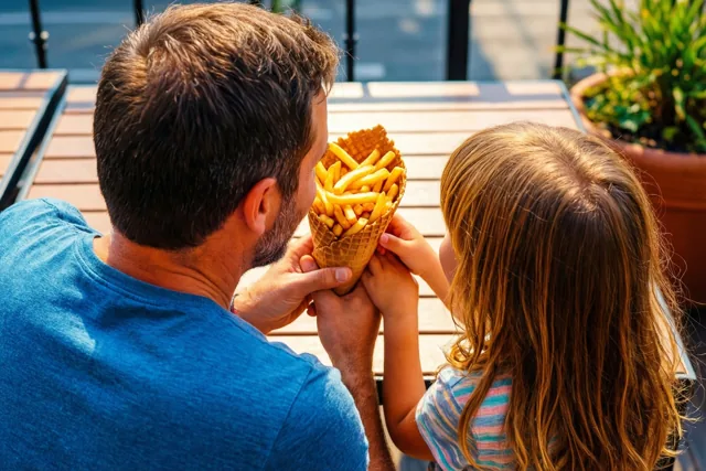 Een vader en kind delen van achteren gezien een wafelhoorntje gevuld met frieten op een houten terras in warm zonlicht.