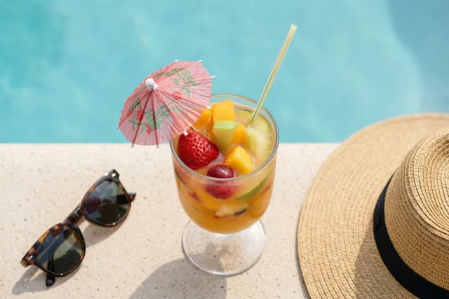 Een glas fruitcocktail met een papieren parasolletje en een rietje staat naast een zonnebril en strohoed op een lichte ondergrond bij een zwembad.