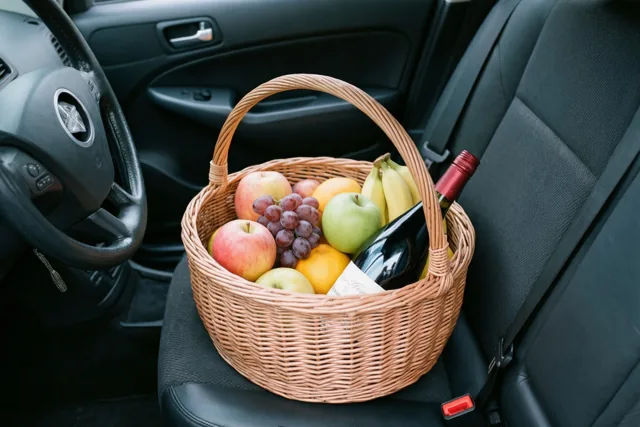 Een rieten mand met fruit en een fles wijn staat op de passagiersstoel van een auto met een donkergrijs interieur.