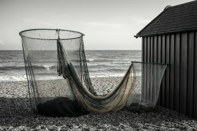 Een groot en een kleiner garnalennet leunen tegen een houten strandcabine op een kiezelstrand bij zee, weergegeven in zwart-wit.