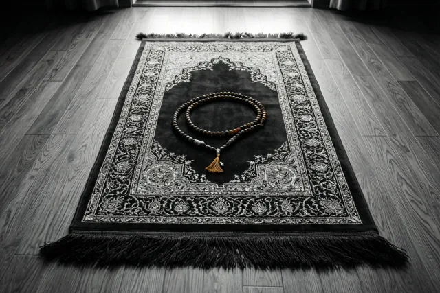 Een gebedskleed met fijne patronen en een snoer gebedskralen ligt op een houten vloer in het ochtendlicht uit een deuropening, in een monochroom beeld met warme tinten op de kralen.