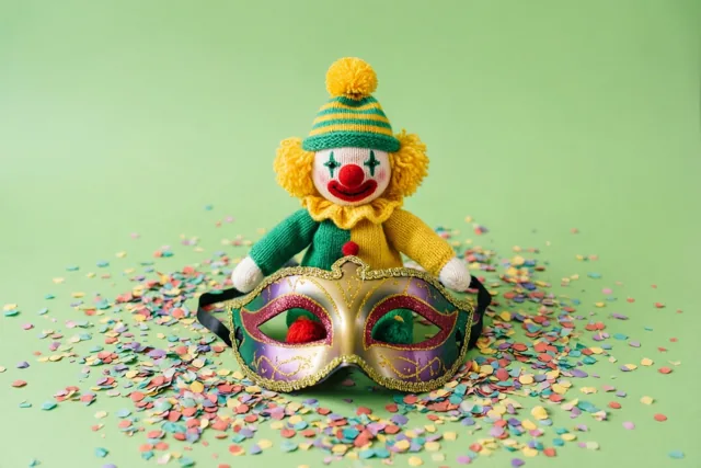 Een klein gebreid clownspopje met een gestreepte pomponmuts zit op een glinsterend carnavalsmasker tussen kleurrijke confetti tegen een effen groene achtergrond.