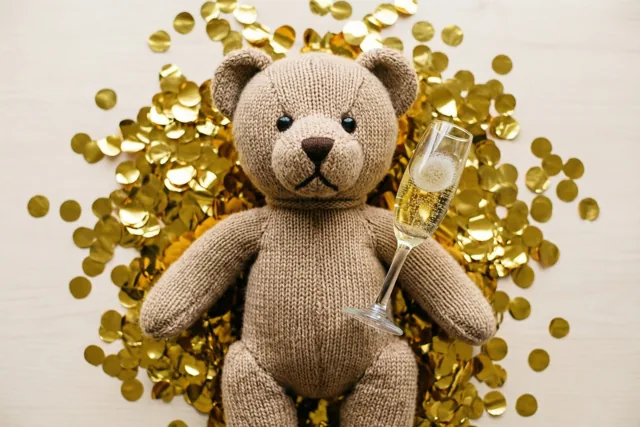 Een gebreide teddybeer zit rechtop met een miniatuurchampagneglas op een licht houten ondergrond waarop gouden confetti ligt.