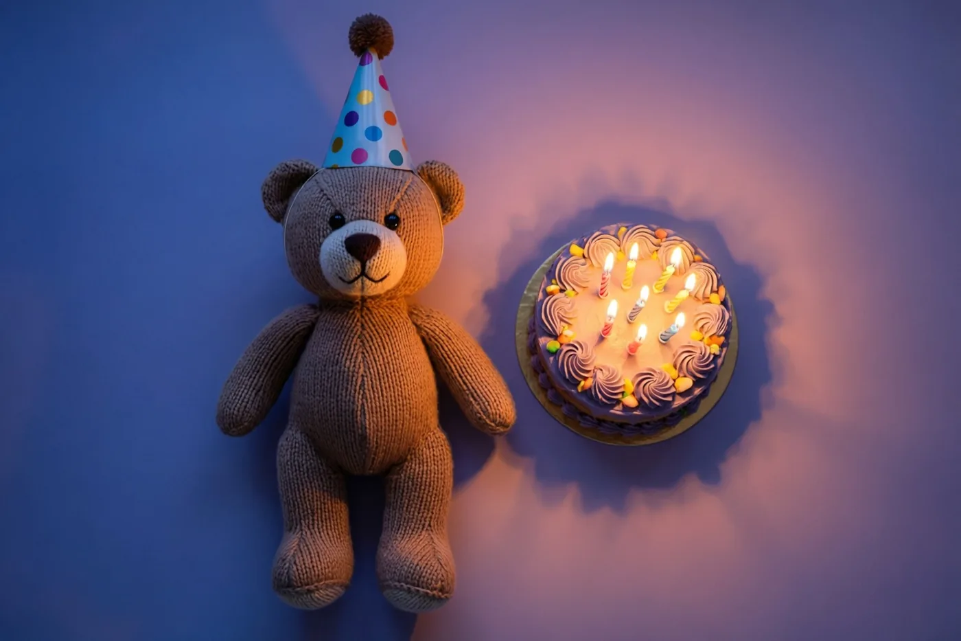 Een gebreide teddybeer met een kleurrijk gestippeld feesthoedje staat naast een verjaardagstaartje met brandende kaarsjes tegen een zacht verlichte blauw-paarse achtergrond.