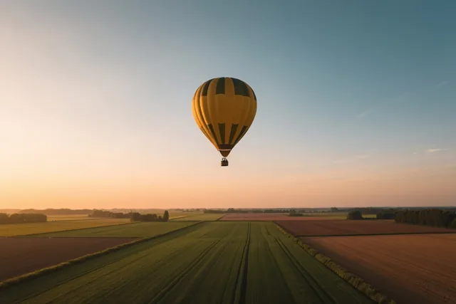 Een geel-zwart gestreepte luchtballon zweeft boven groene en bruine akkers onder een avondhemel die van oranje naar blauw verloopt.