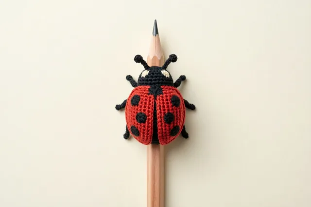 Een rood gehaakt lieveheersbeestje met zwarte stippen zit op een geslepen houten potlood tegen een effen gebroken witte achtergrond.