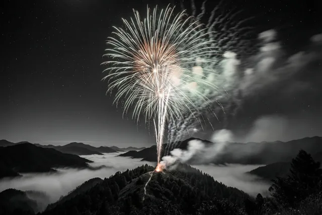 Gekleurd vuurwerk barst 's nachts los boven een bergrug en een mistige vallei in een verder monochroom landschap met sterke contrasten.