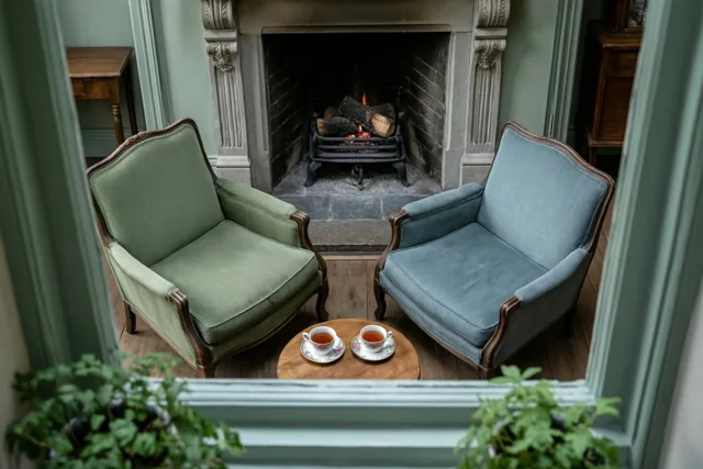 Door een lichtgroen raamkozijn zijn een groene en een blauwe oude fauteuil te zien bij een houten tafeltje met twee kopjes thee voor een brandende haard, met planten op de voorgrond.