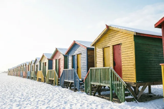 Gele, rode, blauwe en groene houten strandcabines staan met sneeuw op hun daken op een besneeuwd strand onder een bleke hemel.