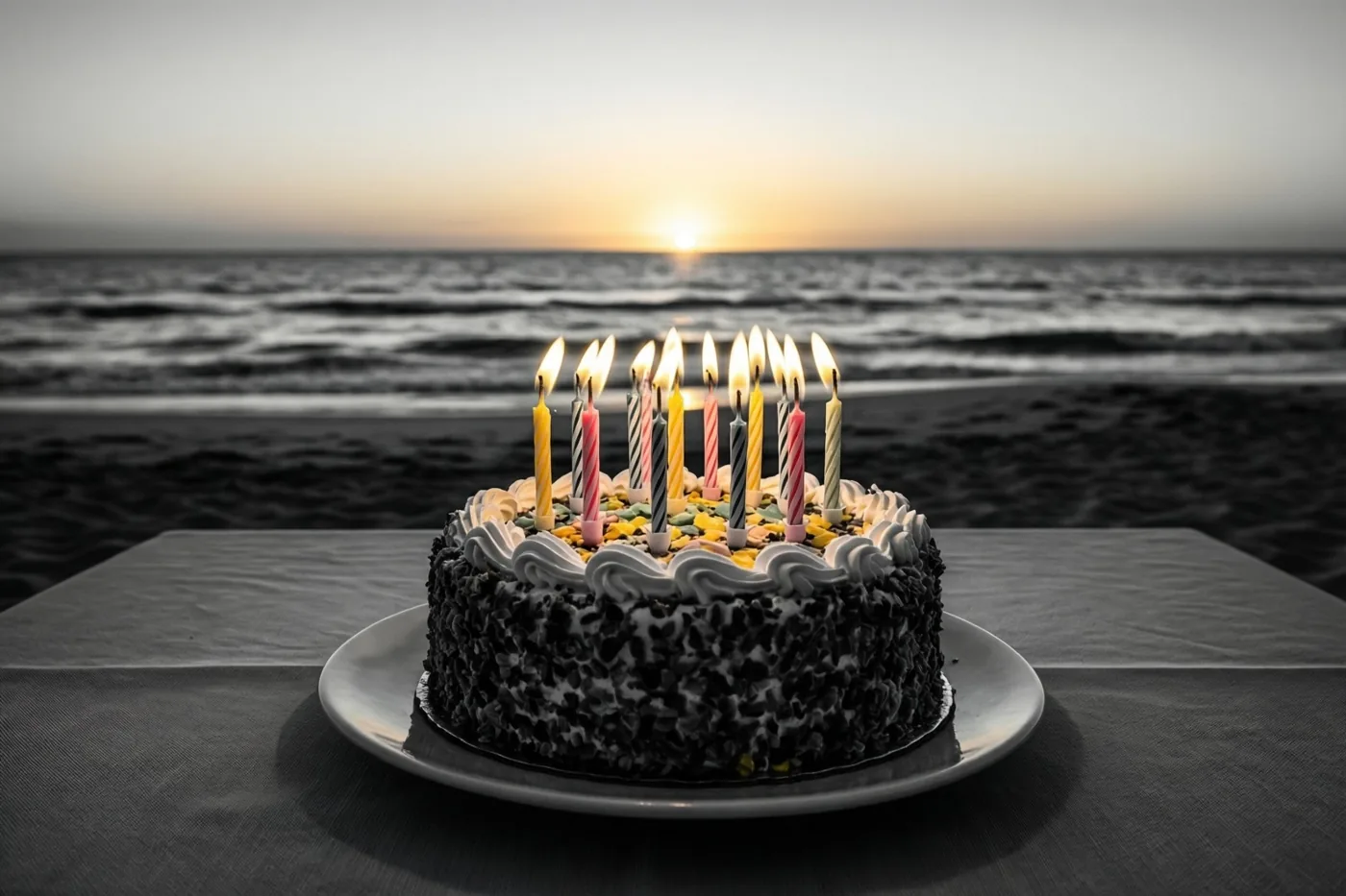 Een verjaardagstaart met brandende kaarsjes staat op een tafel op het strand bij zonsondergang, waarbij de taart en kaarsjes in kleur afsteken tegen de vrijwel kleurloze zee en achtergrond.
