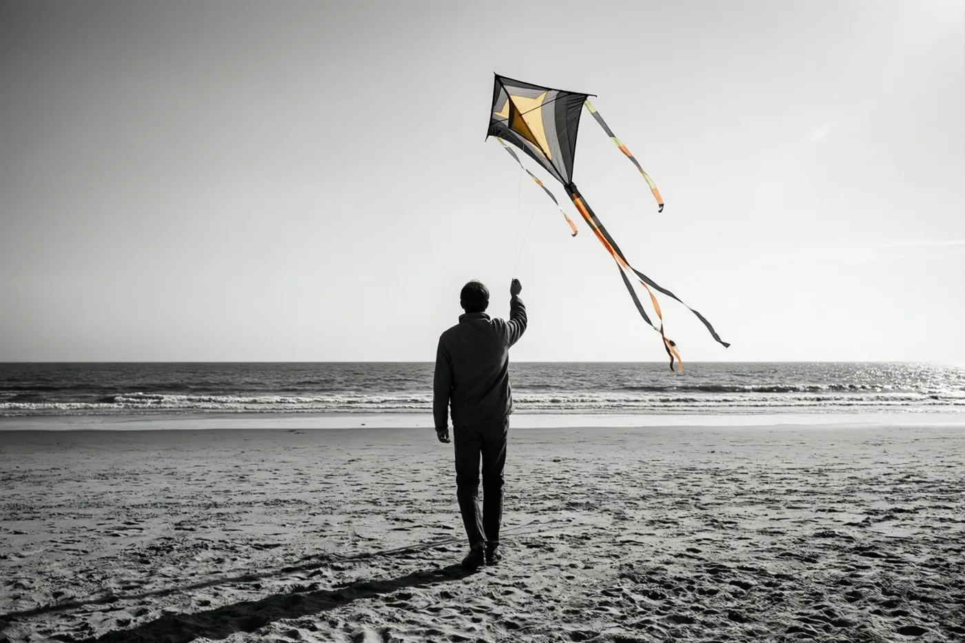 Een persoon staat met de rug naar de kijker op een breed zandstrand en laat een vlieger met lange gekleurde staarten op, waarbij alleen de vlieger kleur heeft in het zwart-witbeeld.