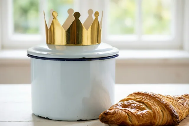 Een witte emaillen broodtrommel met een gouden papieren kroon op het deksel staat op een houten oppervlak naast een goudbruine croissant, met een helder raam op de achtergrond.