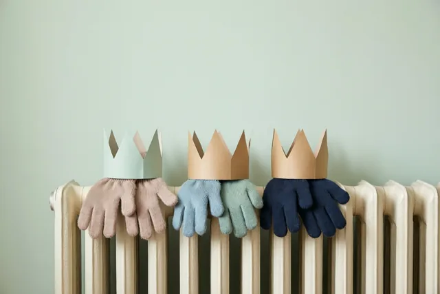 Drie paar kinderwanten met elk een papieren kroon liggen op een witte radiator tegen een lichtgroene muur, in zachte pastelkleuren.