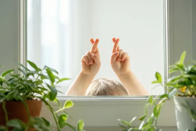 Achter een raam zijn kinderhanden met gekruiste vingers te zien, omringd door potplanten en zacht natuurlijk licht.