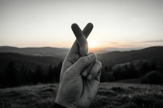 Een opgeheven hand met gekruiste vingers staat voor glooiende heuvels in zwart-wit met een warme gloed van de zonsopgang op de achtergrond.