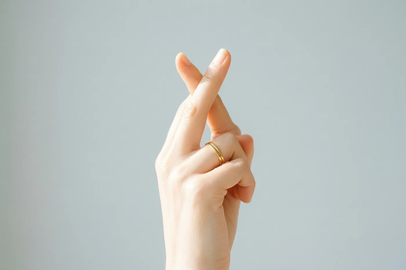 Een hand met gekruiste vingers en een fijne gouden ring is te zien tegen een zachtgrijze achtergrond.